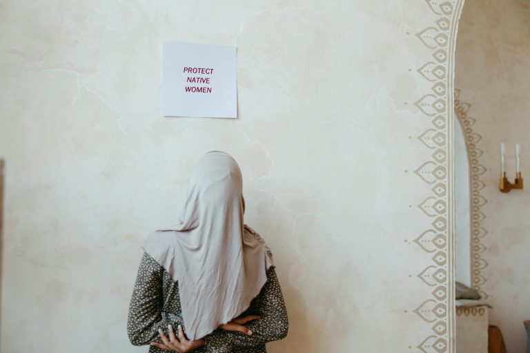 A woman in a hijab stands facing a wall with a sign reading 'Protect Native Women'.