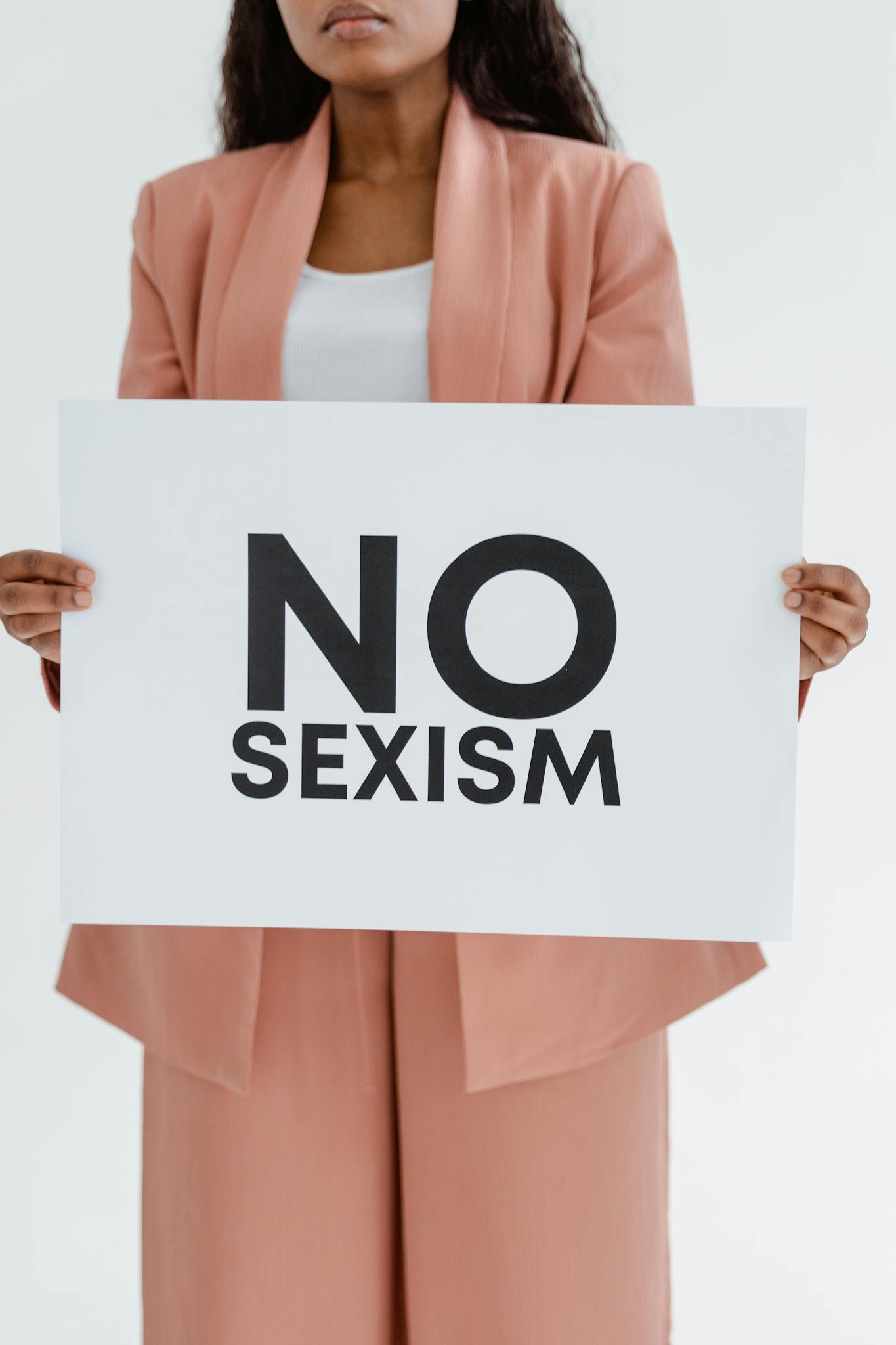 Confident woman stands in feminist pink attire holding a 'No Sexism' sign, advocating equality.