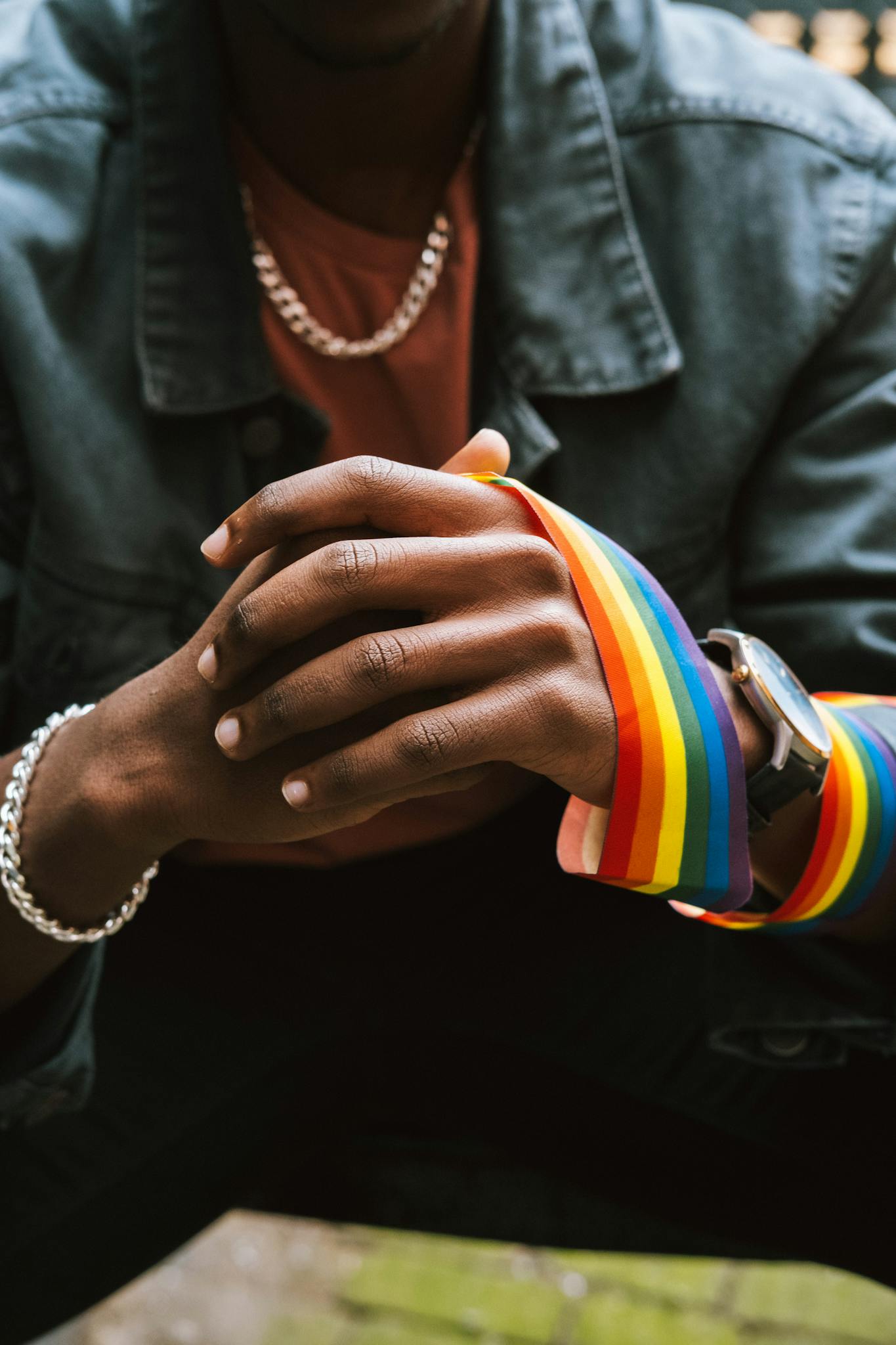Crop anonymous African American homosexual male wearing street style clothes and colorful LGBT ribbon on arm while sitting on street with hands clasped