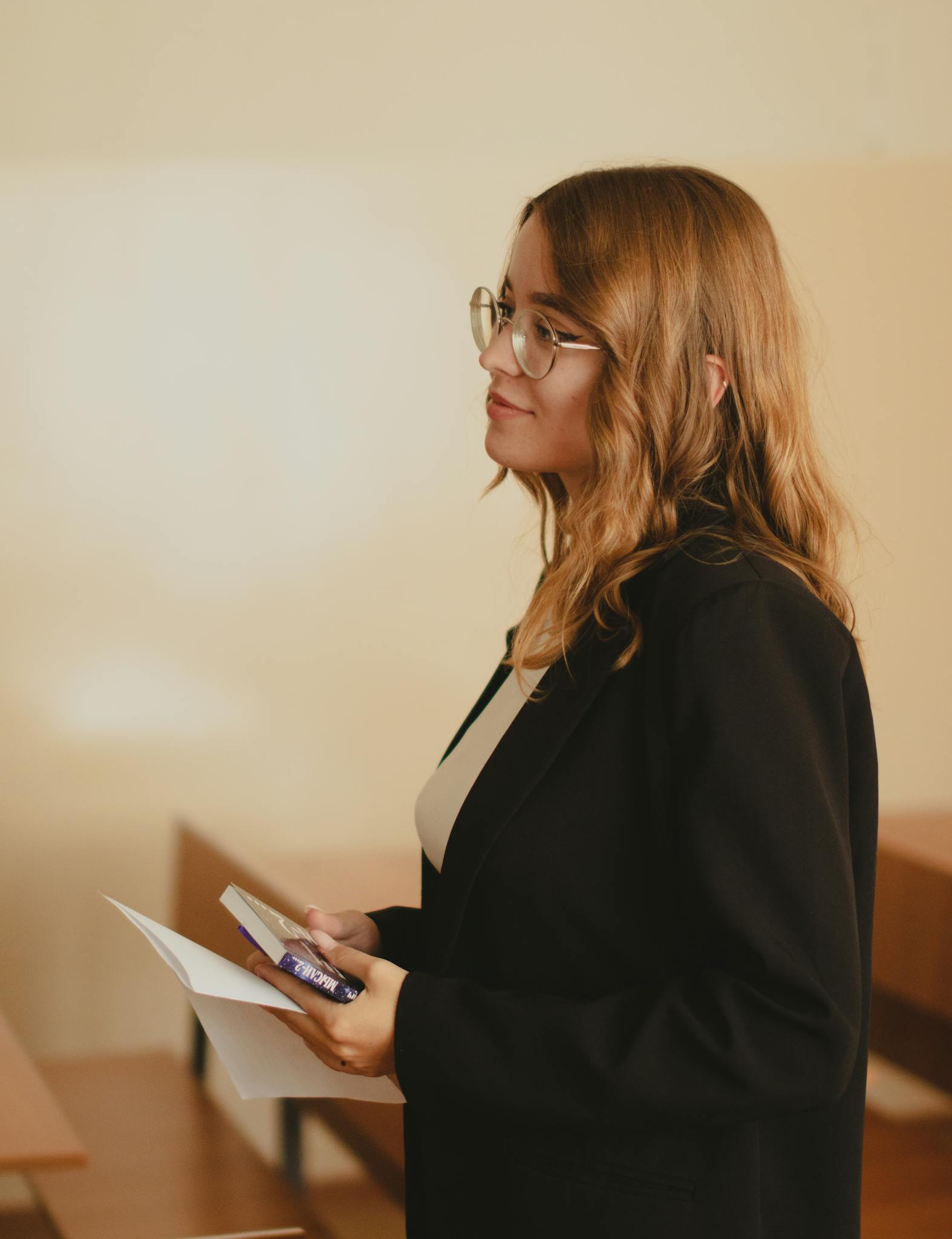 Profile of a young woman in glasses holding papers in a classroom setting.
